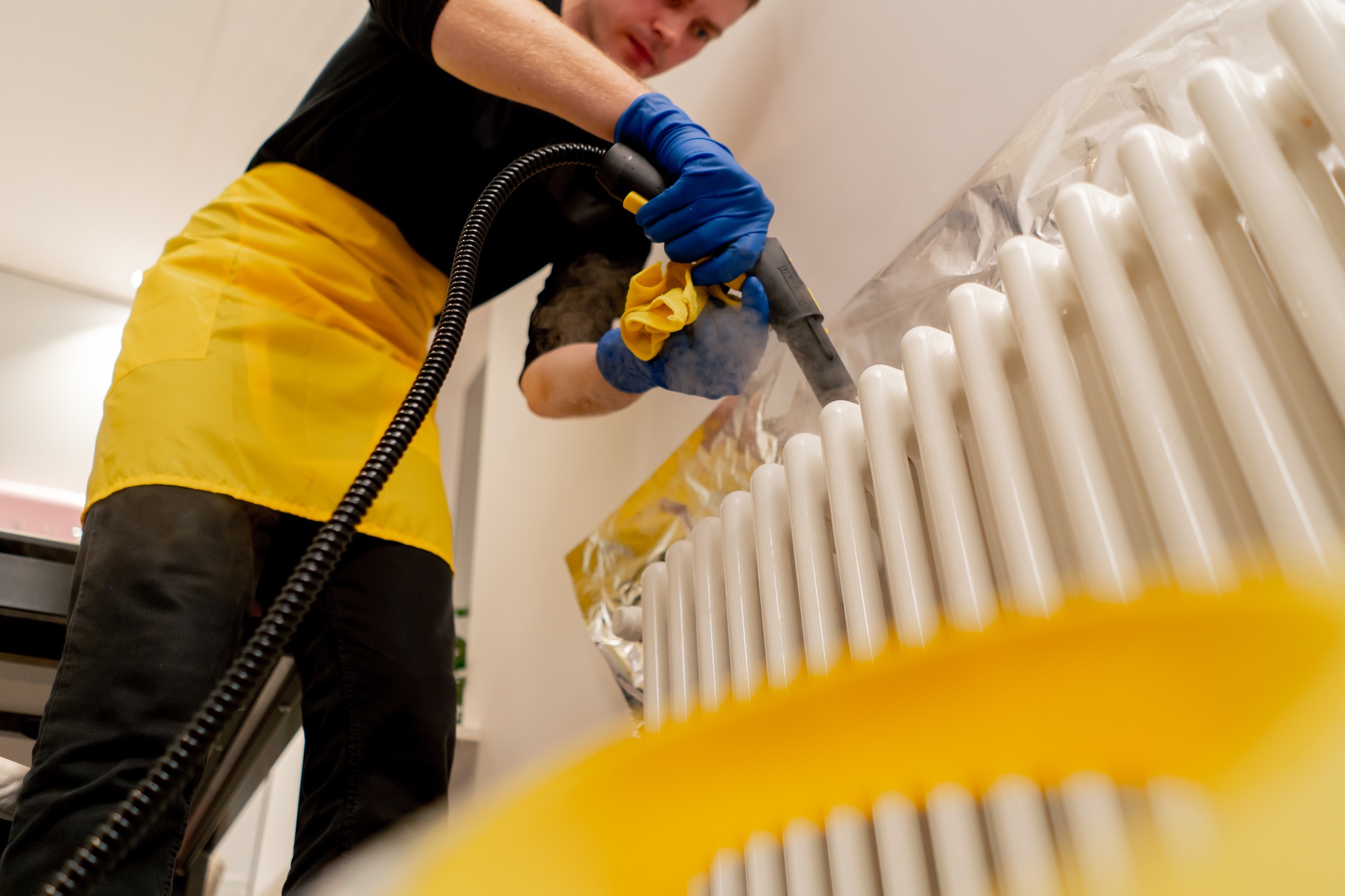 low shot in an apartment cleaner in a yellow apron washes a white radiator with a steam generator